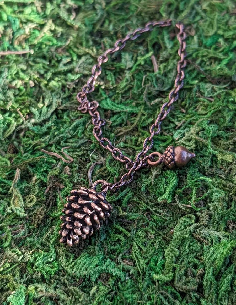 Copper pinecone pendulum with an acorn bead on a copper chain lying on green moss