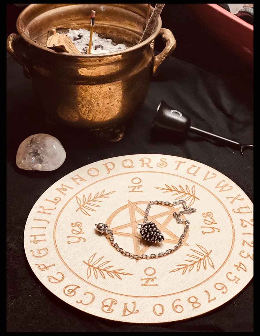 Silver pinecone pendulum on a wooden pendulum board beside a brass cauldron and a quartz stone